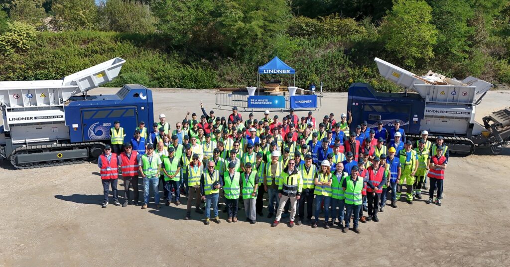 Aerial group photo of around a hundred participants in safety vests standing between a Lindner Urraco ECO 70 DT and a Urraco EVO 75 DT shredder at the Lindner site