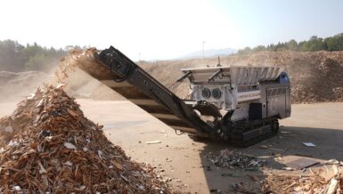 Tracked Lindner Urraco ECO 70 DT twin-shaft shredder throwing shredded waste wood from its discharge conveyor onto a large pile at a wood recycling yard
