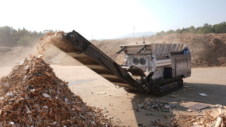 Tracked Lindner Urraco ECO 70 DT twin-shaft shredder throwing shredded waste wood from its discharge conveyor onto a large pile at a wood recycling yard