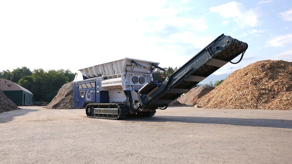 The Lindner Urraco ECO 70 DT mobile shredder on crawler tracks with its long discharge conveyor raised, standing between piles of wood chips on a yard