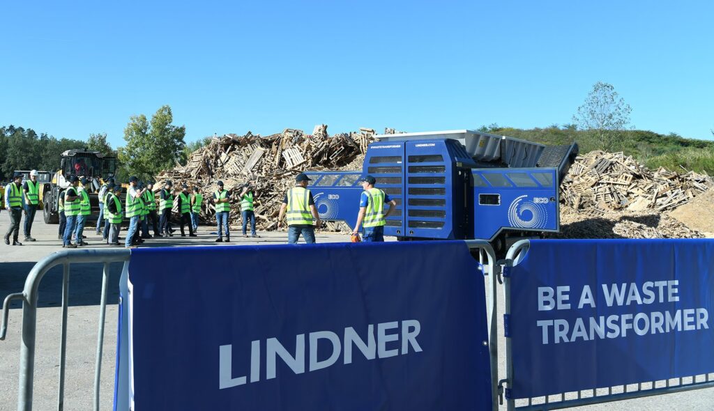 Visitors in high-visibility vests watch a blue Lindner Urraco ECO shredder in front of a large pile of wooden pallets, behind a Lindner banner reading Be a Waste Transformer