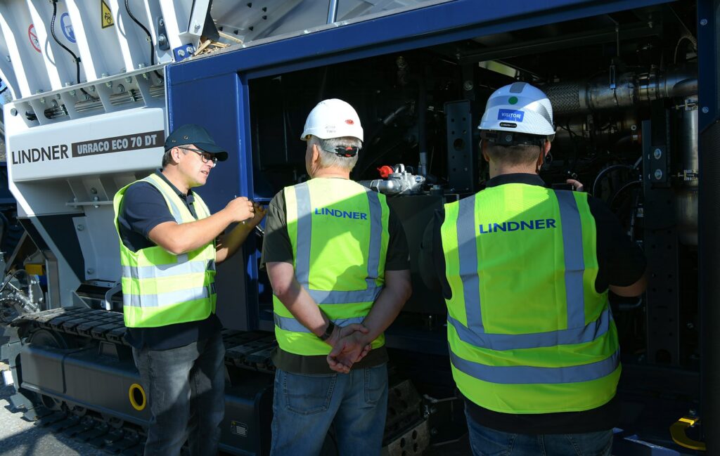 A Lindner employee in a high-visibility vest points out the open engine compartment of the Urraco ECO 70 DT to two visitors wearing Lindner vests and white hard hats