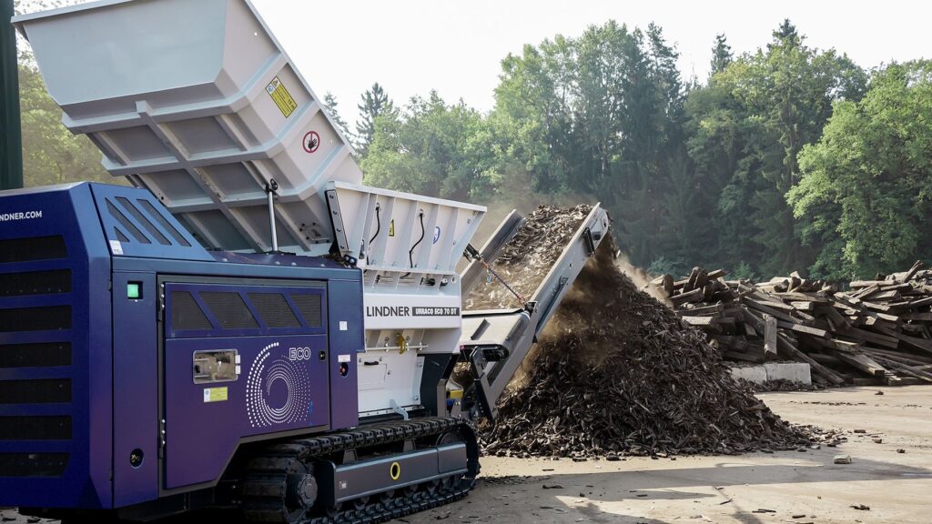 Blue and white Lindner Urraco ECO 70 DT shredder with its hopper extension raised, its conveyor piling dark shredded wood beside a stack of old timber sleepers