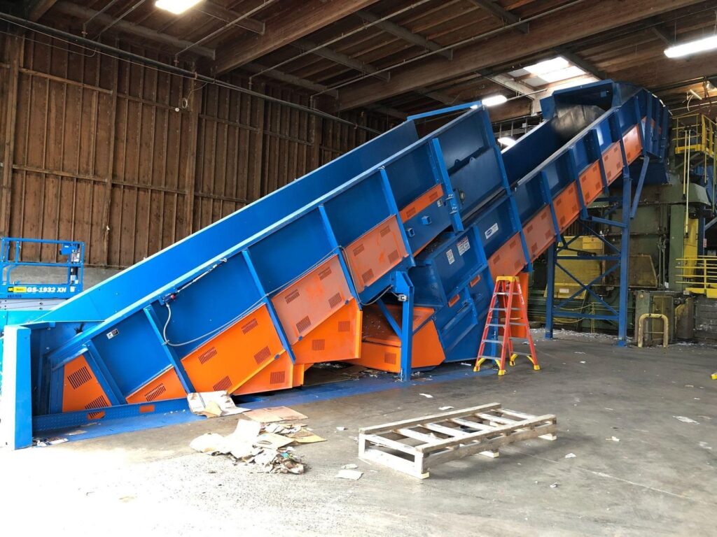 Side view of a Remcon heavy-duty conveyor system with tall blue steel side walls and orange panels on the floor of a paper recycling plant