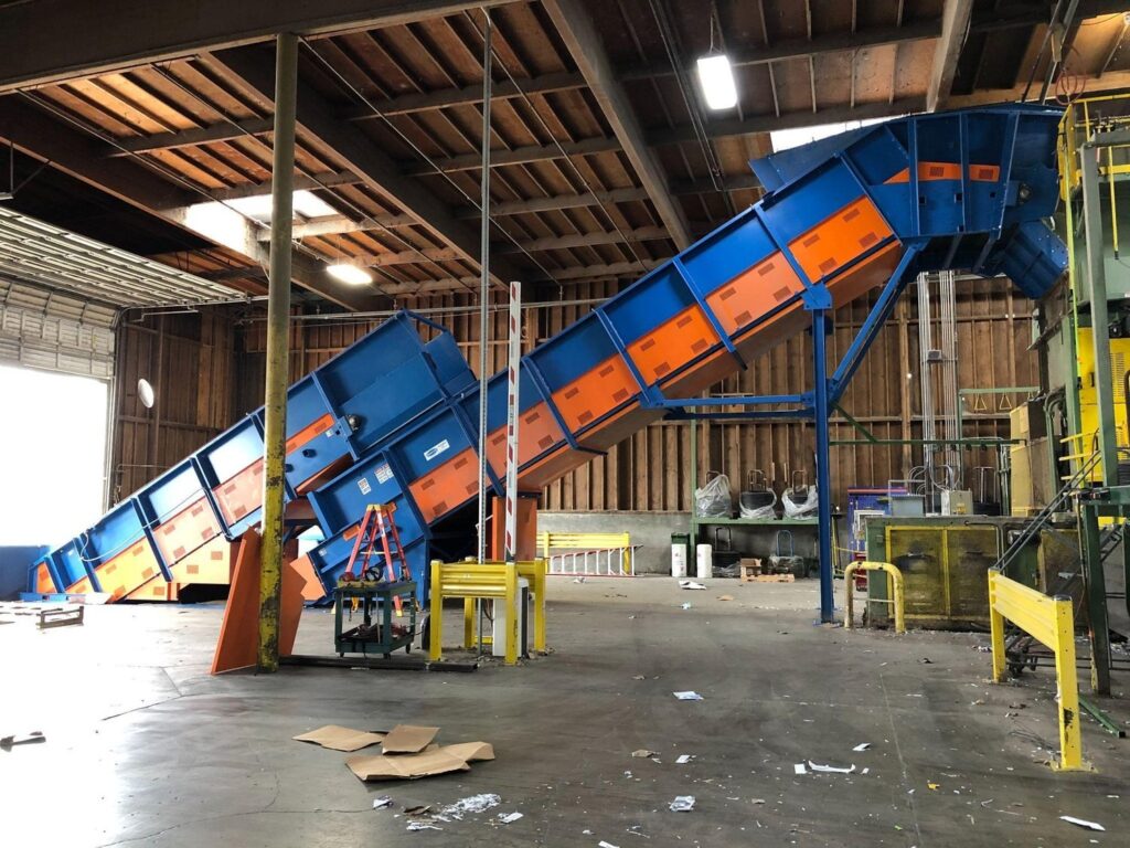Two blue and orange Remcon heavy-duty inclined conveyors rising towards processing equipment inside a paper recycling facility with a timber roof