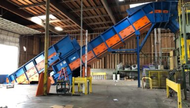 Two blue and orange Remcon heavy-duty inclined conveyors rising towards processing equipment inside a paper recycling facility with a timber roof
