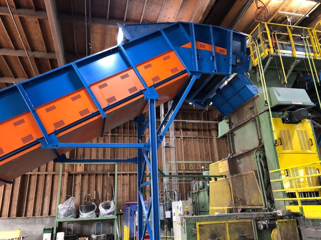Close-up of the discharge end of a blue and orange Remcon inclined conveyor on steel support legs, feeding processing equipment in a recycling plant