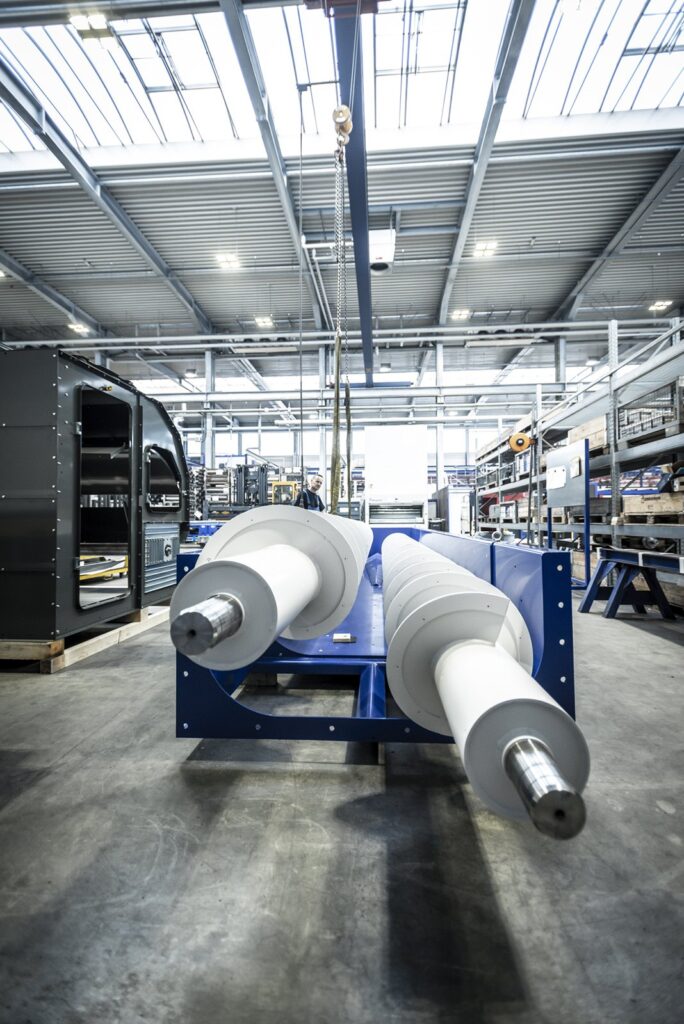 Two large white shafts with steel ends lying in a blue steel frame in a bright production hall, a worker in the background