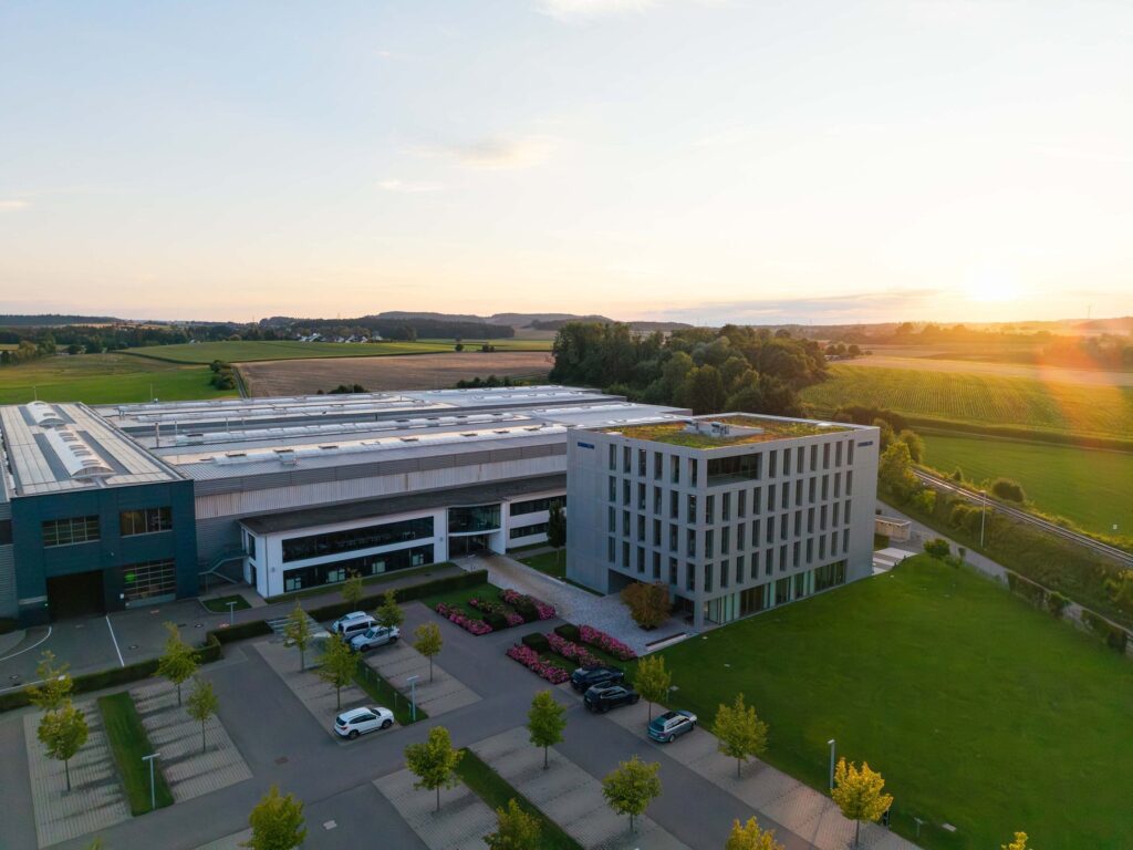Drone view of a large production hall and a modern six-storey office building with green roof, surrounded by fields at sunset