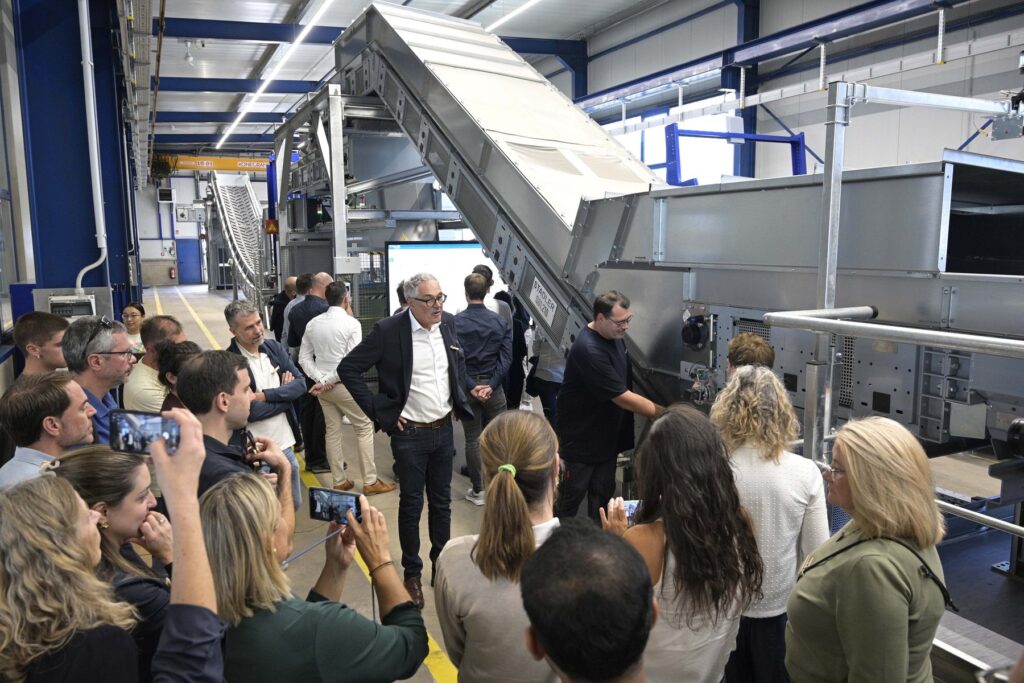 Group of Summit participants, several filming with phones, gathered around Willi Stadler next to an inclined conveyor in the Test Center