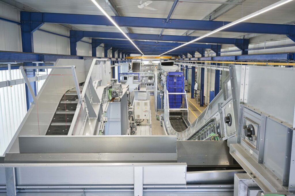 Long hall with blue steel beams and a sequence of galvanized conveyors, sifters and switch cabinets forming a sorting line