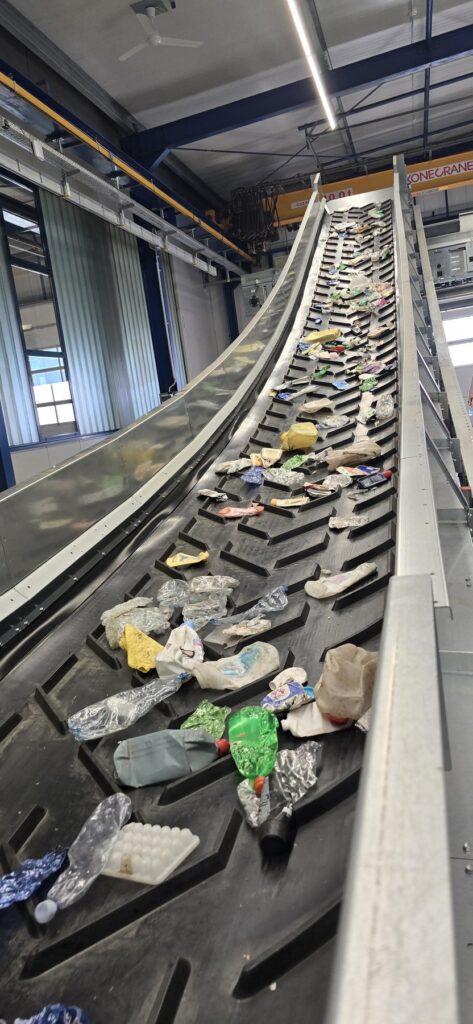 Steep black chevron conveyor belt carrying crushed plastic bottles, film and packaging upwards in a test hall