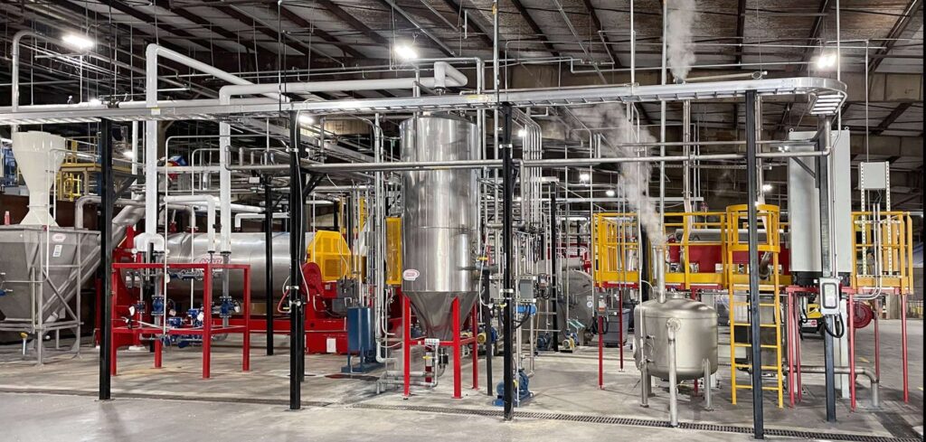 Overview of an AMUT plastic recycling plant inside a factory hall, with red machine frames, a large stainless steel tank, yellow safety platforms and steam rising from the process equipment