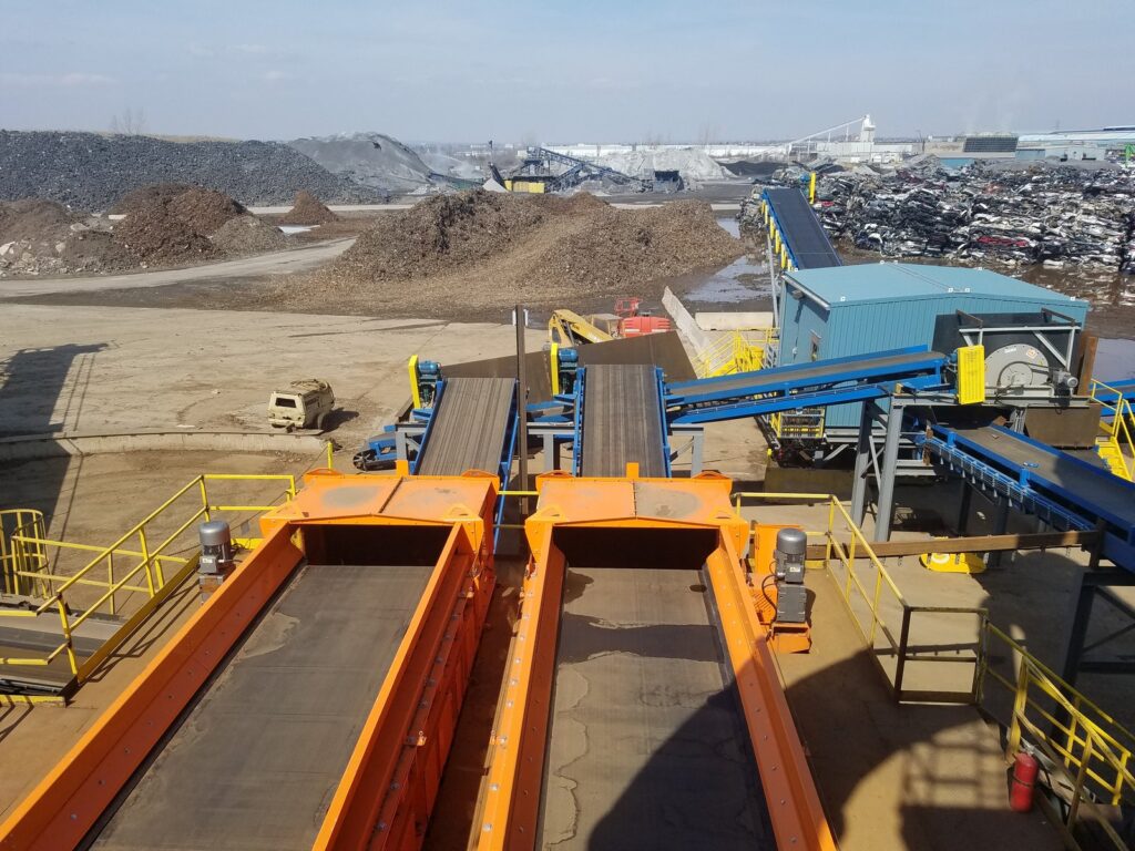 Two orange Eriez Shred1 Ballistic Separator vibratory feeders in the foreground with blue conveyors leading to a magnetic scrap drum, in front of a scrap yard with piles of shredded material and crushed cars