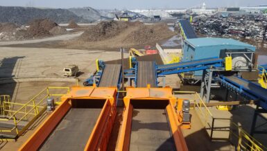 Two orange Eriez Shred1 Ballistic Separator vibratory feeders in the foreground with blue conveyors leading to a magnetic scrap drum, in front of a scrap yard with piles of shredded material and crushed cars