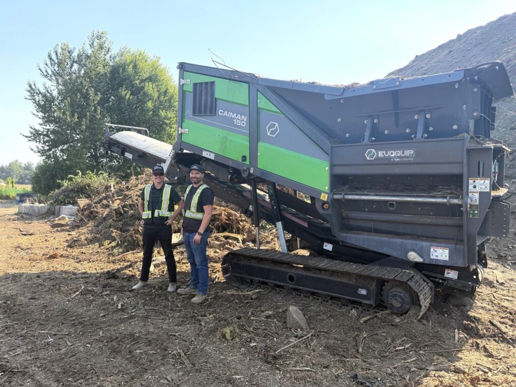 Two men in high-visibility vests stand beside a green and grey EvoQuip Caiman 150 tracked twin-shaft shredder next to a pile of wood and green waste