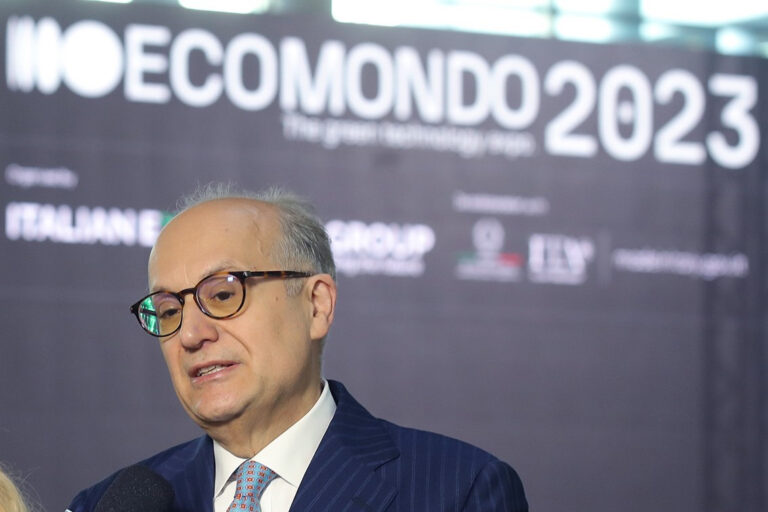 Man in a pinstripe suit and glasses speaking into a television microphone in front of an Ecomondo backdrop