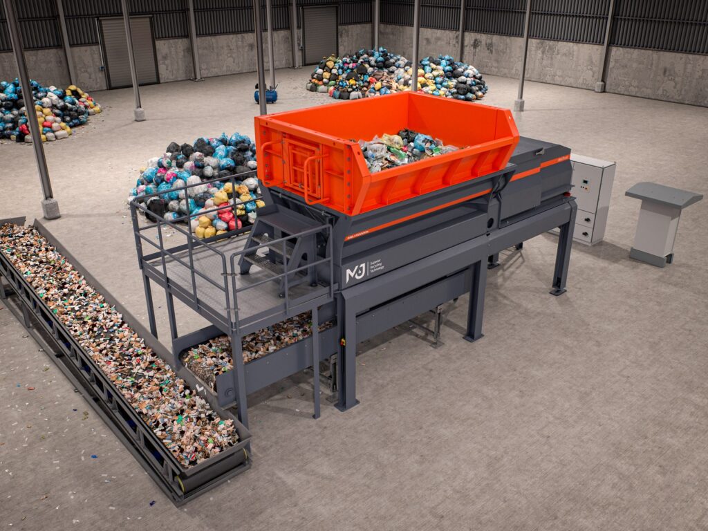 Aerial view of the orange M&J P2000e single-shaft electric pre-shredder with feed hopper, platform and discharge conveyor in a waste treatment hall with piles of household waste bags