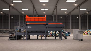 Side view of the M&J P2000e electric pre-shredder with orange feed hopper, UltraBoost drive section, discharge conveyor and control cabinets, with piles of waste bags behind the machine