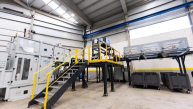 Industrial hall with a Girbau Sortech feeding unit on the left, a raised PICVISA ECOSORT optical sorter on a yellow-railed platform in the centre and grey collection bins on the right