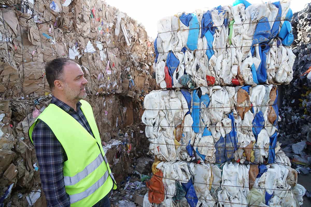 Man in a high-visibility vest looking at stacked bales of compressed plastic canisters next to cardboard bales