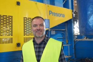 Smiling man in a high-visibility vest standing in front of a yellow and blue Presona baler