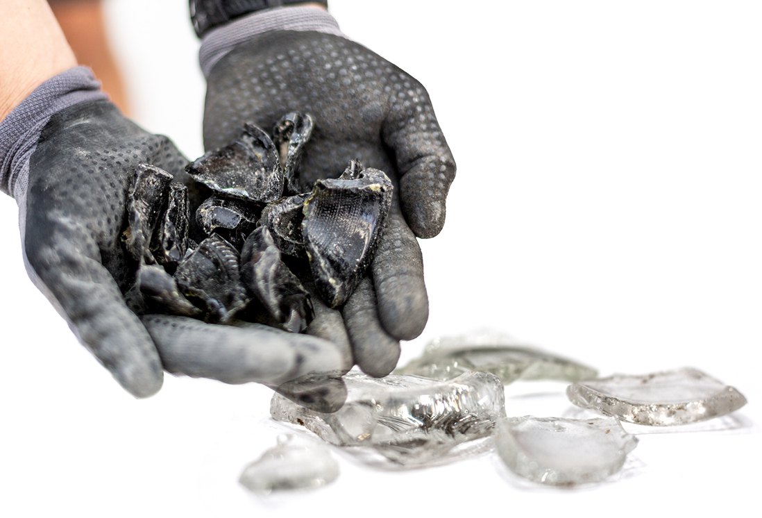 Gloved hands holding dark glass cullet above pieces of clear glass