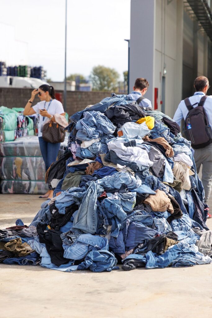Tall heap of used jeans and other garments on a concrete yard, with bales of textiles and visitors in the background