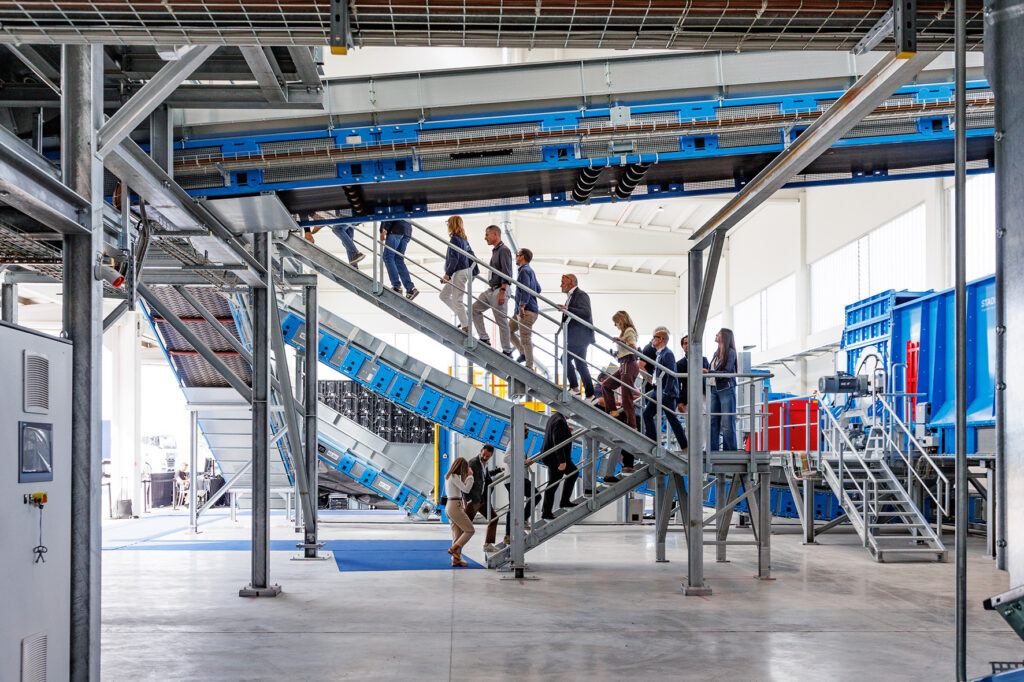 Line of visitors walking up a galvanized steel staircase beside inclined blue conveyors in a bright industrial hall