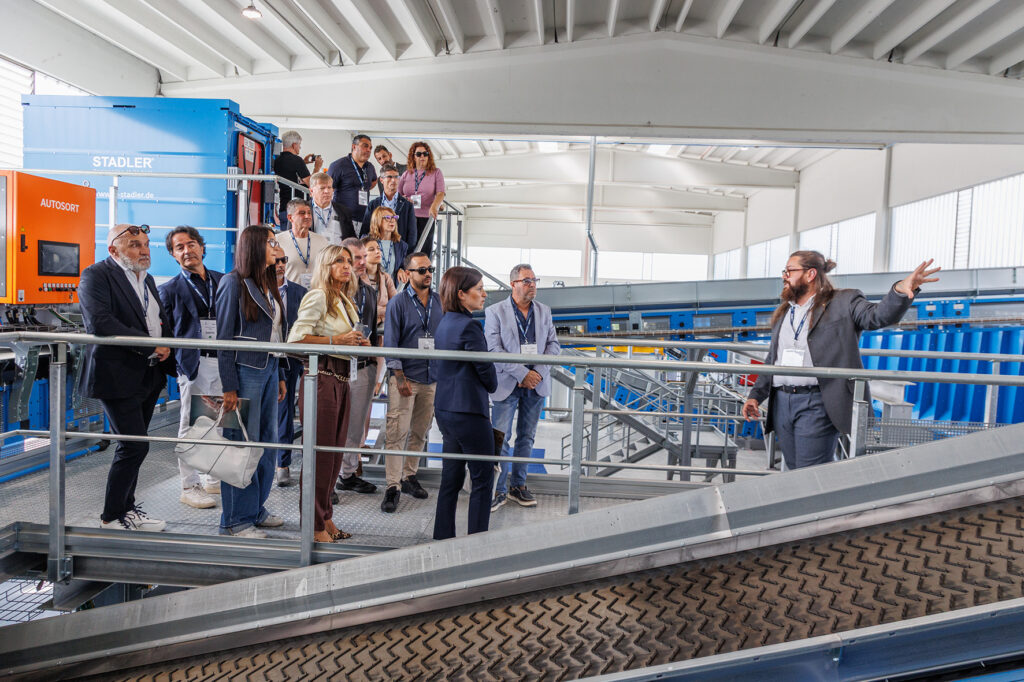 Guide gesturing to a group of visitors on a galvanized steel platform next to an orange AUTOSORT control cabinet and a blue STADLER unit, with a chevron conveyor belt in the foreground
