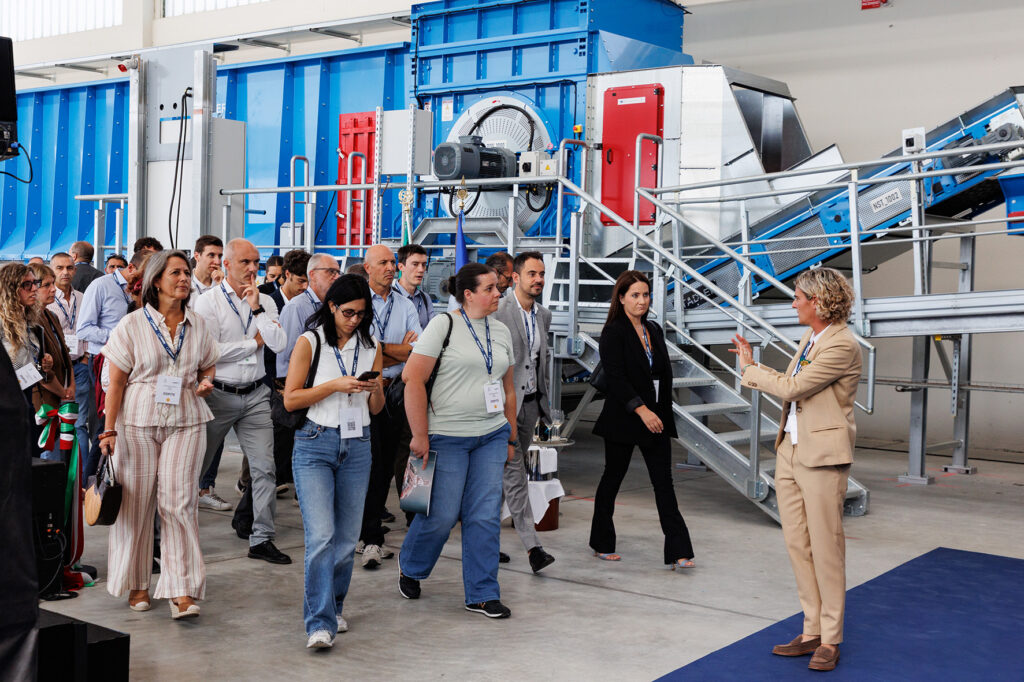 Guide in a beige suit addressing a group of visitors with lanyards in front of a blue drum machine and an inclined conveyor