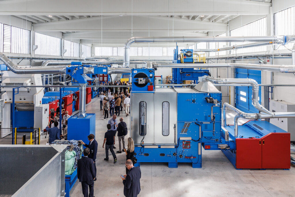 High-angle view of a bright hall with blue and red textile processing machines, silver air ducts and groups of visitors on the floor
