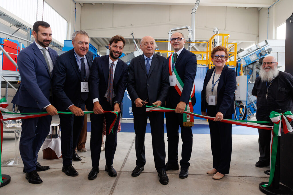 Group of officials in suits holding an Italian tricolour ribbon as it is cut, with blue and yellow plant equipment behind them