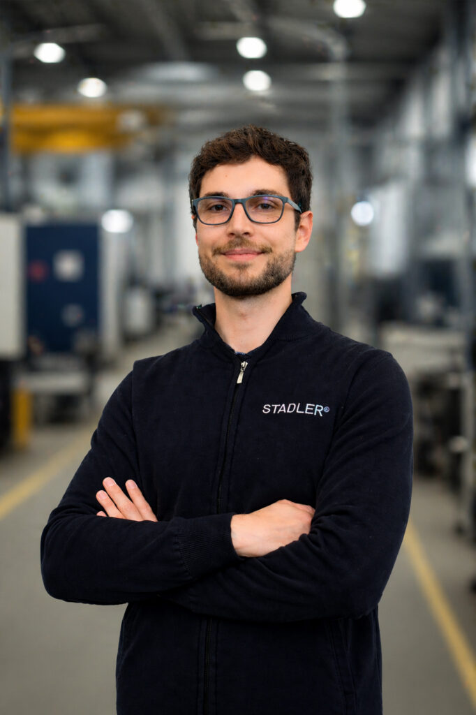 Man with glasses and a beard wearing a dark jacket with the STADLER logo, standing with arms crossed in a production hall
