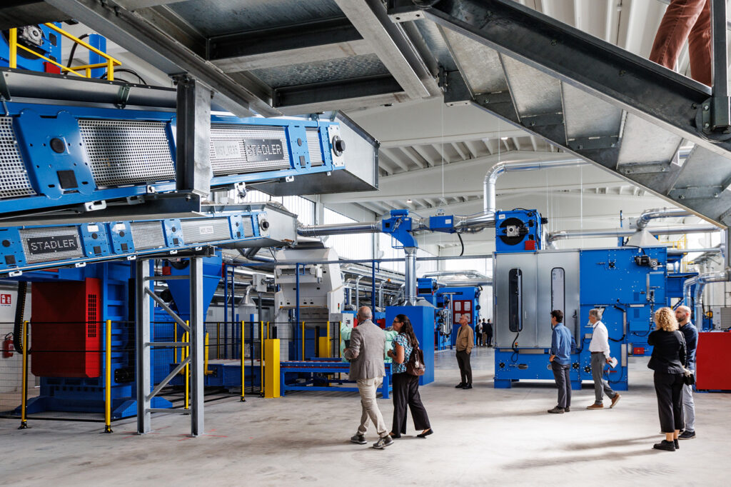 Blue STADLER conveyors mounted under a steel platform in a bright hall, with visitors walking between blue and red textile processing machines