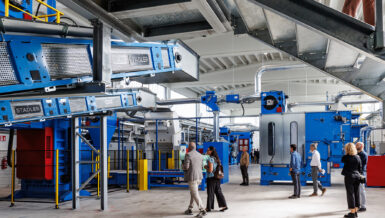 Blue STADLER conveyors mounted under a steel platform in a bright hall, with visitors walking between blue and red textile processing machines