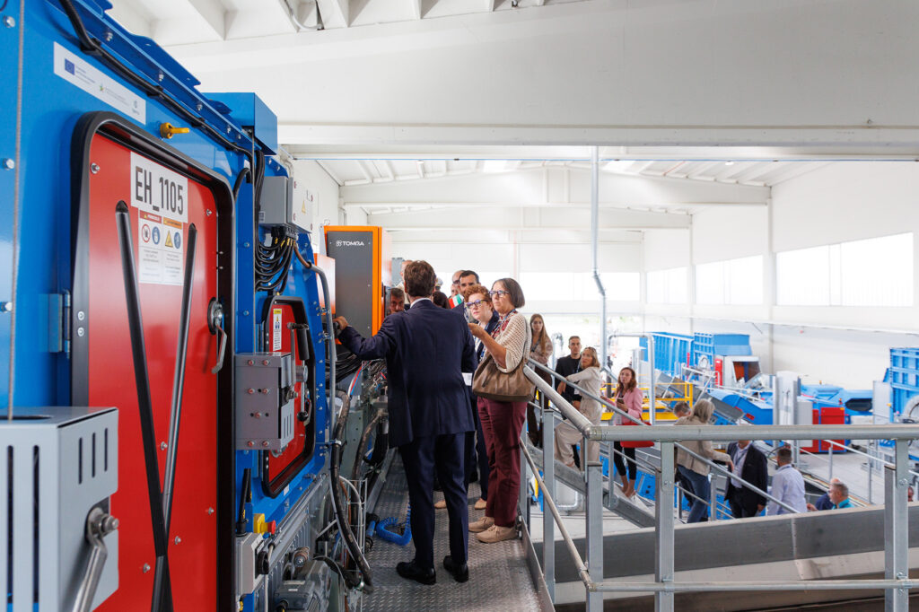 Close-up of a blue and red machine housing with an orange TOMRA unit behind it, while visitors stand on the walkway alongside