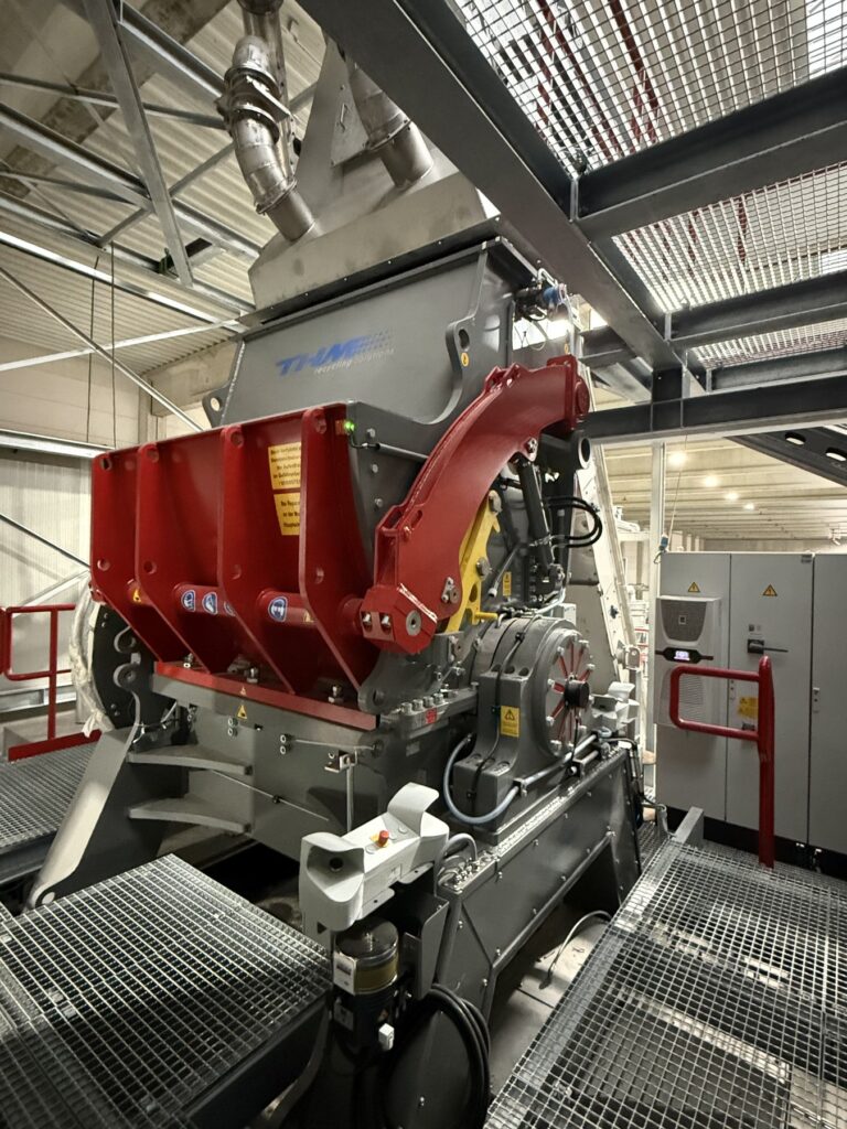 Grey THM granulator with red hydraulic housing arms and feed hopper on a steel grating platform, with control cabinets in the background