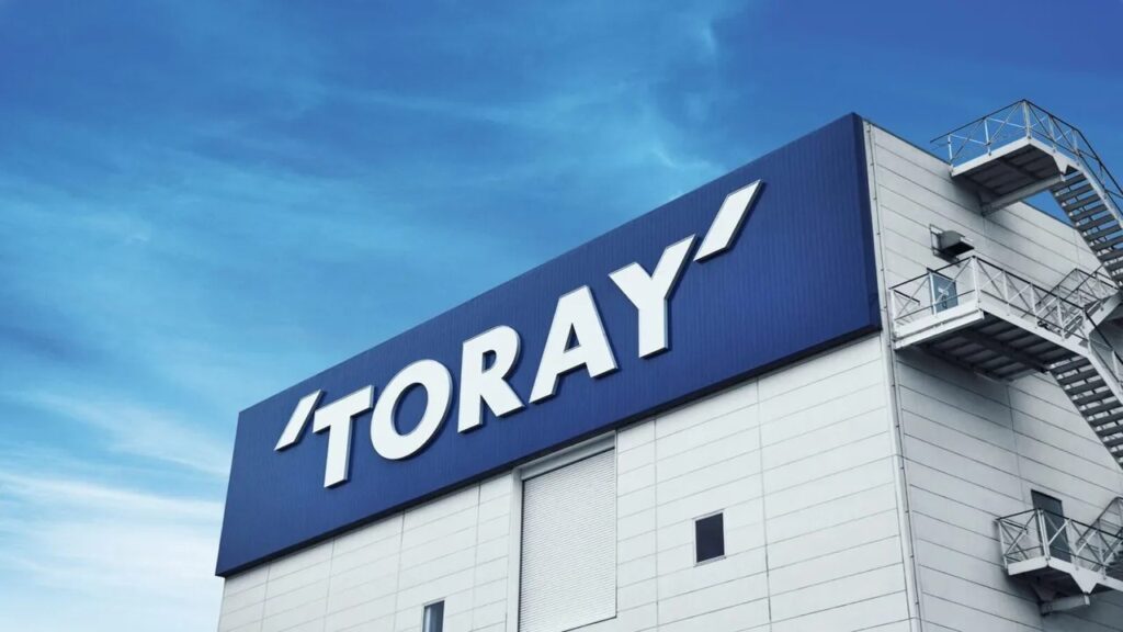 White industrial building with a large blue Toray sign on the roof edge and an exterior fire escape staircase against a blue sky