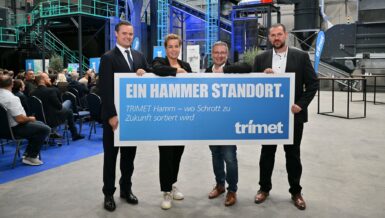 Four people stand on a stage holding a blue banner reading "Ein Hammer Standort" at the opening of the TRIMET aluminum scrap sorting facility in Hamm, with the sorting plant behind them and an audience seated on the left