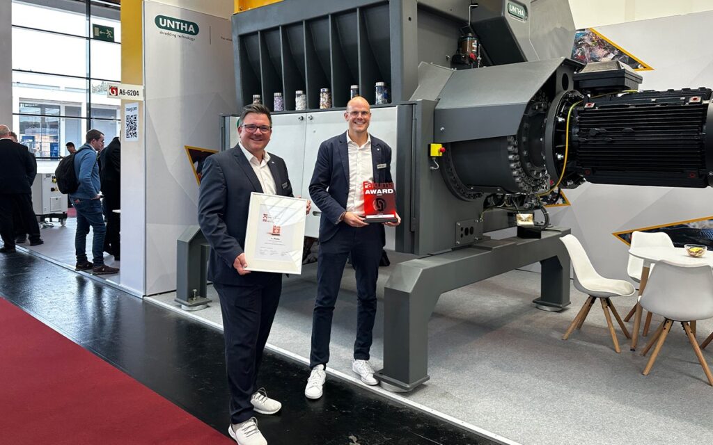 Two men holding the FAKUMA Innovation Award certificate and trophy, standing in front of a large grey UNTHA MR 1800C PRO shredder on a trade fair stand