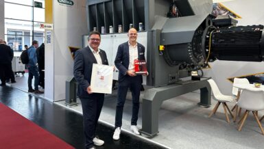 Two men holding the FAKUMA Innovation Award certificate and trophy, standing in front of a large grey UNTHA MR 1800C PRO shredder on a trade fair stand