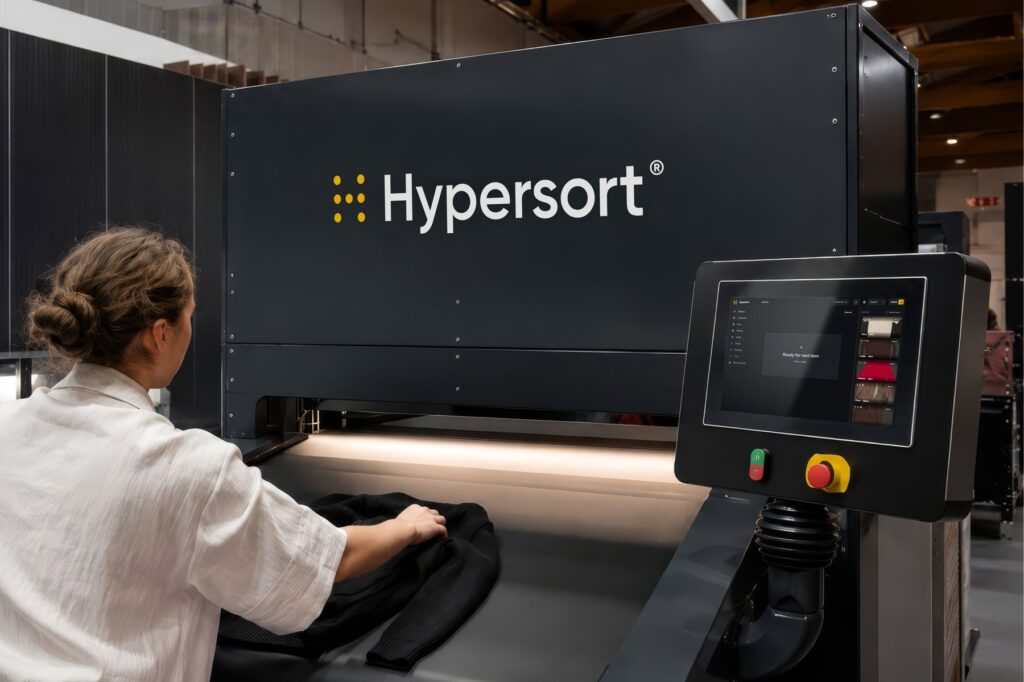 Operator placing a black knitted garment on the conveyor of the Valvan Hypersort machine, with the touchscreen showing the next item ready
