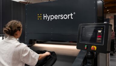 Operator placing a black knitted garment on the conveyor of the Valvan Hypersort machine, with the touchscreen showing the next item ready